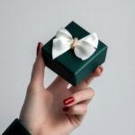 A hand with painted red nails holds out an open dark green gift box in the shape...