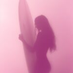 A blurry pink photograph of a woman's silhouette holding up her surfboard, foggy...