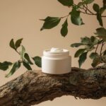 A white cosmetic jar with cream on top, resting on an old tree branch and surrou...