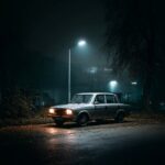 Ein altes silbernes Auto steht auf einem nassen Asphaltplatz, umgeben von Dunkel...