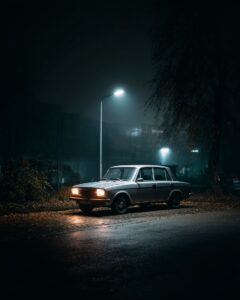 Ein altes silbernes Auto steht auf einem nassen Asphaltplatz, umgeben von Dunkel...