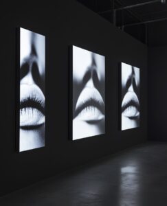 A black wall with three vertical video screens showing lips and eyes in the dark...