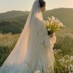 A woman in a white wedding dress holding calla lilies, with a flowing cape, stan...