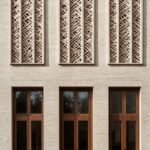 A minimalist building facade made of beige brick with large wooden windows and a...