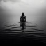 Black white dark blue brown image of dark skinned man standing on infinite water...