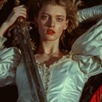 Epic scene, art shows a woman lying on a bed, holding a large sword across her c...