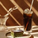 A realistic advertising photo of black ice coffee and green tea cake, in a glass...