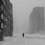 A solitary figure walks down a snowy, 1990, desolate urban street flanked by som...