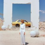 A clad model standing next to a white car, in the style of joel sternfeld, georg...