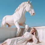 A girl in white is lying under an antique statue of a white horse, in the style ...