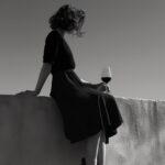 a woman sits on a wall with wine around her feet, in the style of black and whit...