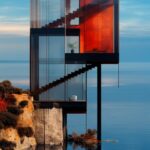 A transparent house on a cliff overlooking the water, has an extra large window ...