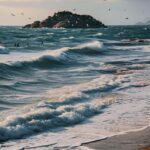 On the Mediterranean seaside, people are walking on the beach. There is a small ...