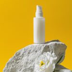 A bottle of white face cream is placed on a white rock with a white flower besid...
