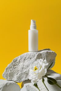 A bottle of white face cream is placed on a white rock with a white flower besid...
